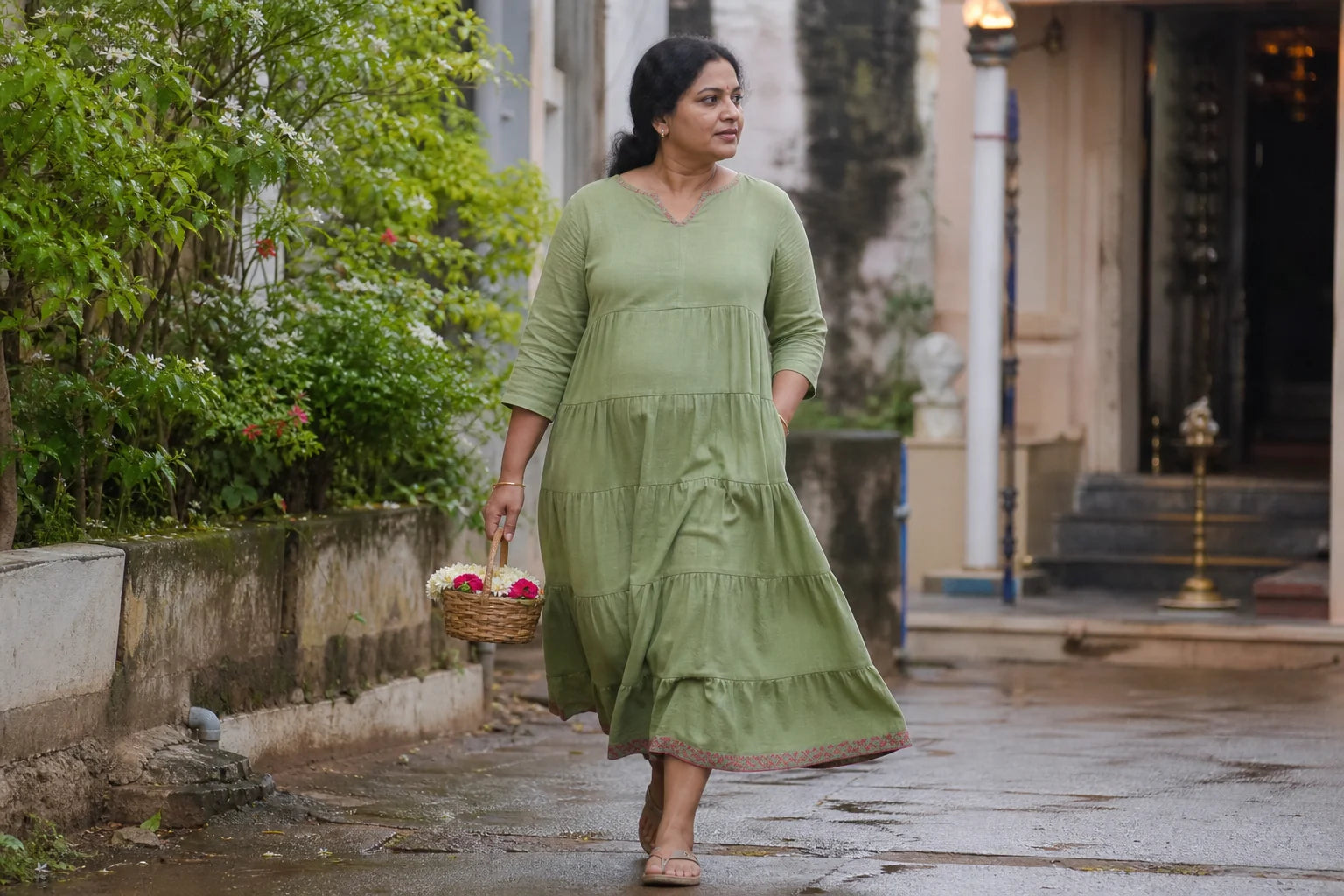 Round-bodied Indian woman carrying flowers in a leaf green tiered day dress near a neighbourhood temple