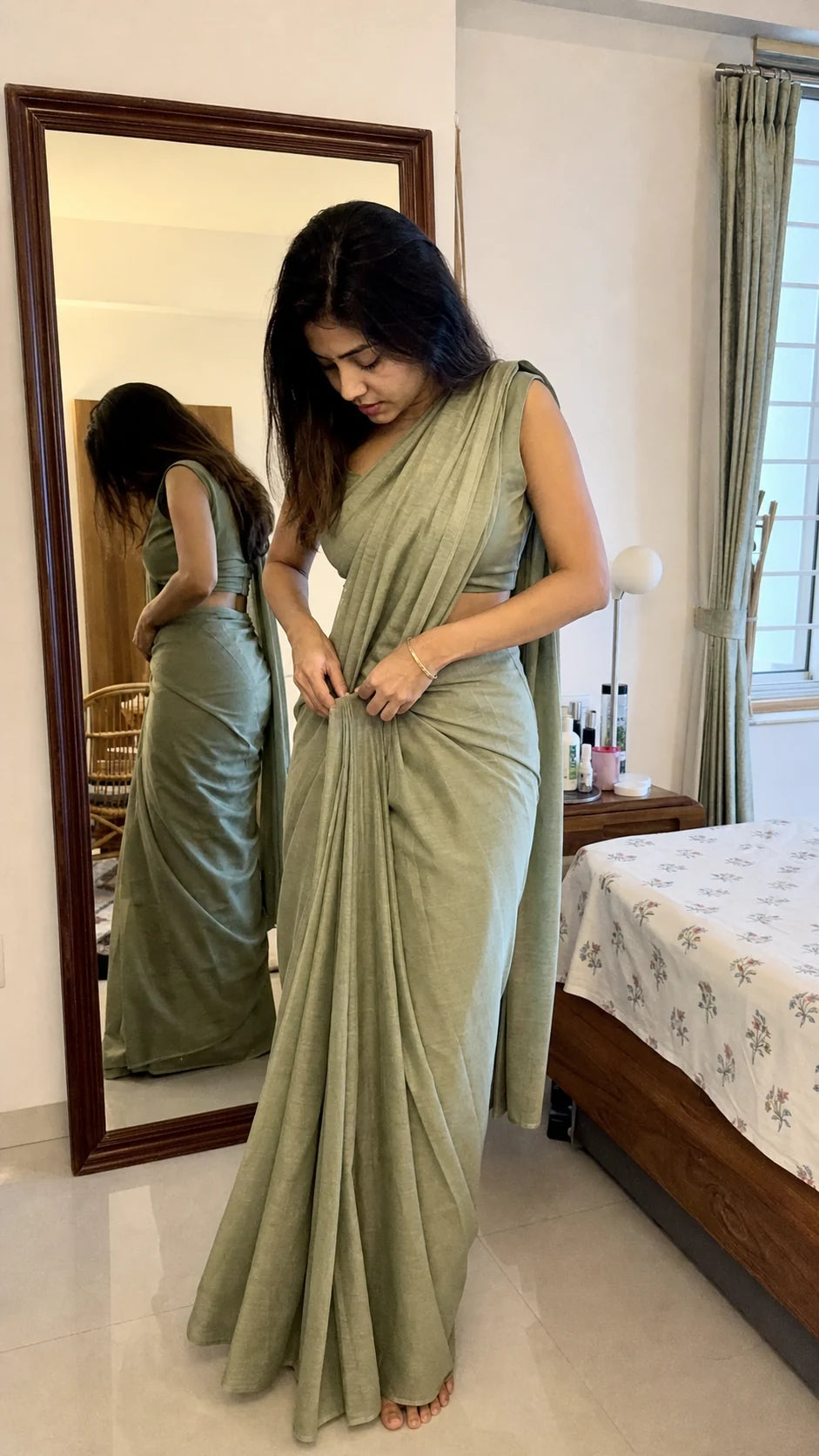 Generated editorial fit study of a petite Indian woman adjusting a muted green saree beside a bedroom mirror